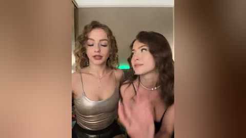 Video of two women, one with curly blonde hair, wearing a metallic top, and the other with straight brown hair, in a pink top, looking at each other in a dimly lit room.