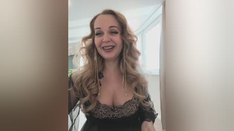 Video of a smiling woman with curly, shoulder-length blonde hair, wearing a black lace bralette and long, gold earrings, standing in a brightly lit room with large windows.