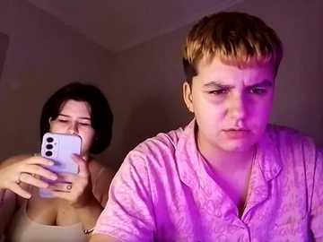 Video of a young woman with short, dark hair taking a selfie, while a young boy with blonde hair and a pink shirt looks disinterested in the background.