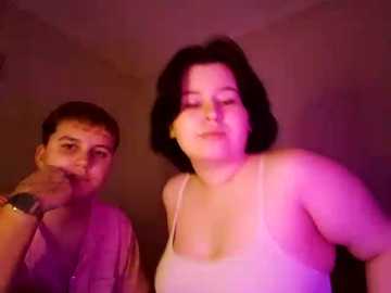 Video of a chubby woman with short black hair in a pink tank top and a man with short brown hair in a grey shirt, both looking towards the camera, dimly lit with a pinkish hue.