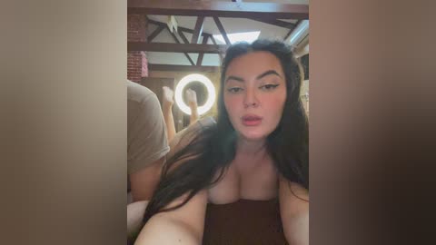 Video of a young woman with long dark hair, wearing a brown top, lying on her stomach in a dimly lit, rustic room with exposed beams and a round mirror.