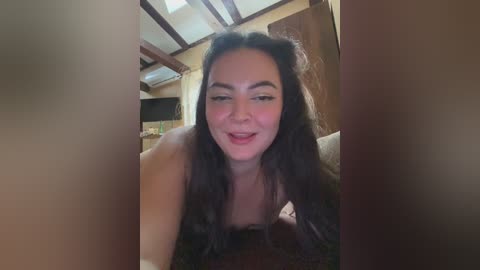 A video of a young, light-skinned woman with long dark hair, smiling, wearing a strapless dark top, in a cozy, rustic room with wooden beams and a flat-screen TV.