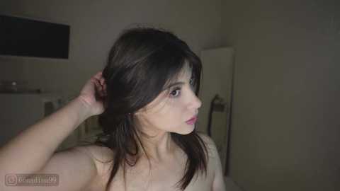 Video of a young, fair-skinned woman with dark hair, standing in a dimly lit room, topless, looking contemplative.