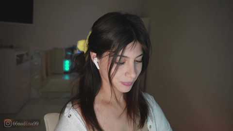 A video of a young woman with long dark hair, wearing a white cardigan and white earbuds, sitting in a dimly lit room with a green light in the background.