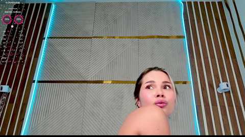 Video of a young woman with light skin, brown hair, and a slender build, captured from below, showing her bare shoulders. She stands in a futuristic room with geometric patterns and glowing blue lights.