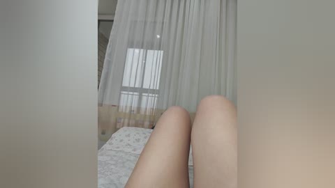 Video of a bedroom with a bed, sheer curtains, and light-colored walls. Legs, likely female, are in the foreground, slightly blurred, and the room is softly lit.