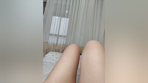 Video of a pair of pale legs on a bed, with sheer white curtains and a floral-patterned quilt in the background.