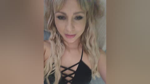 A video of a young Caucasian woman with long, wavy blonde hair, wearing a black lace-up top, smiling. The background is blurred, suggesting a close-up selfie.