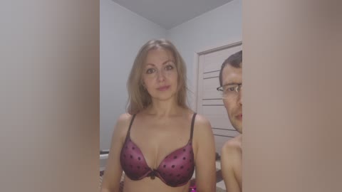 Video of a Caucasian woman with light skin, long blonde hair, wearing a purple polka-dot bra, standing in a dimly lit bedroom with a partially visible mirror reflecting her face.