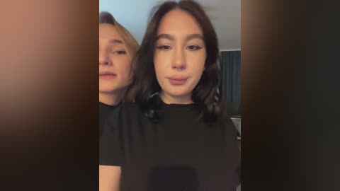 Video of two women, one with short blonde hair and the other with dark, wavy hair, both wearing black tops, standing side by side indoors with a blurred background.