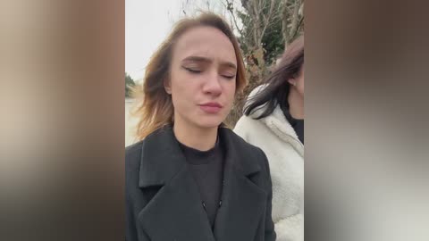 Video of a young woman with fair skin, brown hair, and closed eyes, wearing a black coat over a black turtleneck. Background shows a blurry outdoor scene with trees and foliage.