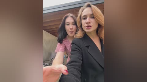 Video of two women standing close together outside, one in a pink patterned top, the other in a black blazer. They both have long hair, and the background features a wooden ceiling and beige wall.