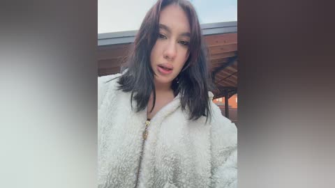 Video of a young woman with shoulder-length dark hair, wearing a fluffy white jacket, standing against a wooden balcony railing.