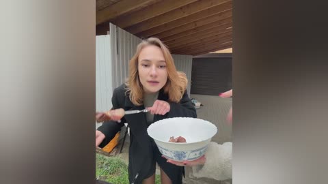 Video of a young Caucasian woman with blonde hair, wearing a black coat, eating ice cream from a blue bowl on a wooden porch.