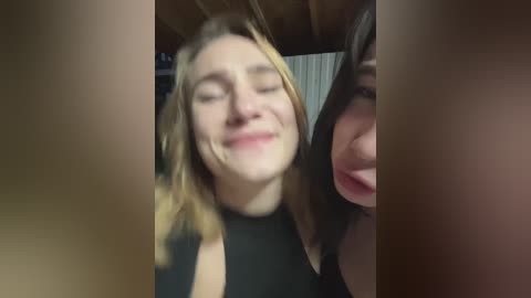 Video of a young woman with shoulder-length blonde hair, wearing a black top, smiling, taken from a selfie angle. Background shows blurred indoor setting with wooden beams and a partially visible person on the right.