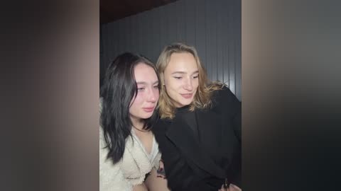 Video of two young women with long hair, one in a white knit sweater, the other in a black jacket, smiling softly at each other. Background is dark and blurry, creating a warm, intimate atmosphere.