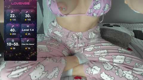 A video of a fair-skinned woman wearing a pink Hello Kitty-themed bra and matching pants, sitting cross-legged on a bed with white and gray bedding. The image includes a game screen with statistics and a \"Level Up\" message.