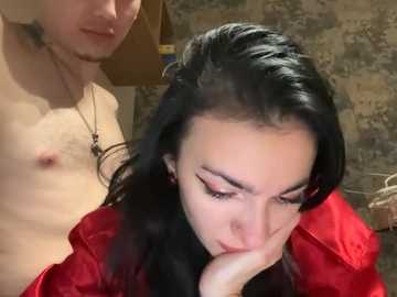 A video of a shirtless, bearded man with fair skin and a tattoo on his chest standing behind a black-haired woman with pale skin, red lipstick, and a red jacket, looking down.