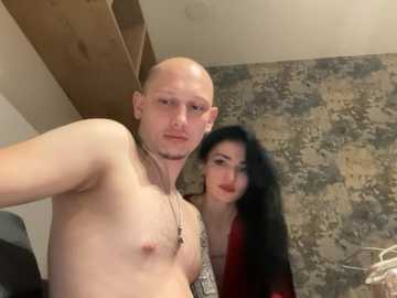 Video of a shirtless, bald Caucasian man with fair skin and a lean build, standing next to a dark-haired woman with a slender figure, both indoors with rustic wooden and stone walls.