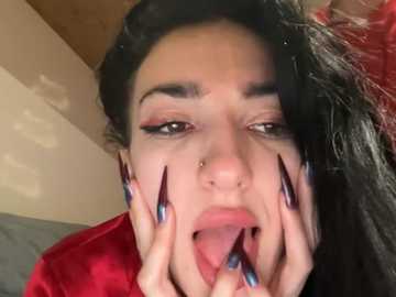 A close-up video of a young woman with long, dark hair, wearing a red velvet top and bold, black eyeliner. She sticks out her tongue and wiggles her long, black-and-blue acrylic nails.