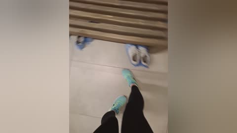 A video showing a person in black pants and turquoise socks standing on a beige tiled floor, with blue painter's tape and a wooden slat wall in the background.