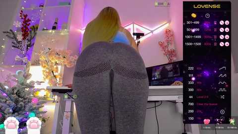 Video of a blonde woman bending over, wearing tight grey leggings, in a brightly lit room with a Christmas tree, digital display, and multiple social media icons.