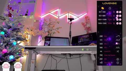 Video of a festive room with a snow-covered Christmas tree, colorful ornaments, and a sleek, minimalist desk with monitors and a microphone. Pink neon \"LOVENSE\" sign and a vibrant, purple-lit background.