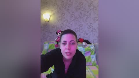 A video of a woman with dark hair and brown eyes, wearing a black shirt, lying on a bed with green and yellow checkered sheets, surrounded by plush toys in a room with floral wallpaper.