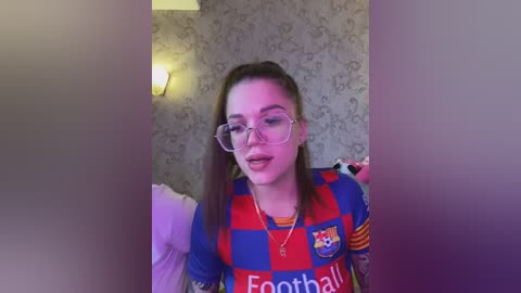 Video of a young woman with light skin, brown hair in a high ponytail, wearing oversized glasses and a Barcelona soccer jersey, indoors with wallpaper and a lamp in the background.