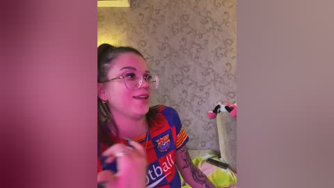 Video of a young woman with light skin and glasses, wearing a red and blue soccer jersey, laughing, with a floral wallpaper background.