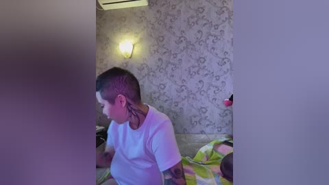 Video of a young boy with short hair and a black tattoo sleeve on his arm, sitting on a bed with a green and yellow patterned blanket. The room has a vintage wallpaper with floral designs and a wall-mounted light fixture.