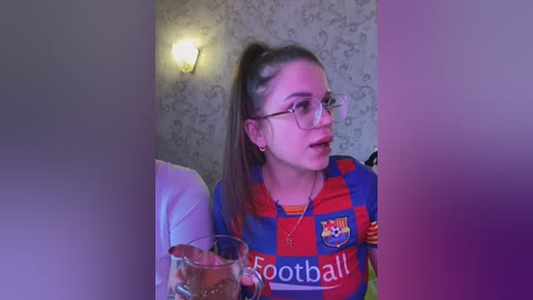A video of a young woman with glasses, brown hair in a high ponytail, wearing a Barcelona soccer jersey, seated at a table with a glass of beer, in a dimly lit, wallpapered room.
