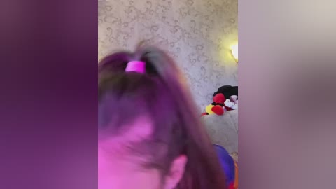 Video of a child with long, dark hair in a high ponytail, wearing a pink headband. The background features a room with a wallpaper pattern, a stuffed duck, and a colorful toy car.