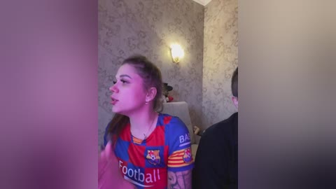 A video of a young woman with light skin and long brown hair, wearing a Barcelona football jersey, standing in a dimly-lit room with floral wallpaper.