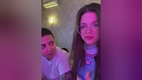 Video of two young people in a dimly lit room. A man with short dark hair and a woman with long brown hair and glasses, both wearing casual clothes. Background features patterned wallpaper and a light fixture.