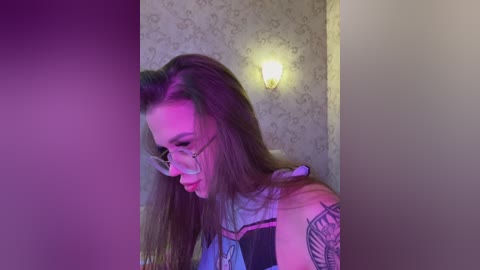 A video of a young woman with long brown hair, wearing glasses, a tattoo on her shoulder, and a dark top, illuminated by purple light. She appears to be in a dimly lit room with floral wallpaper.