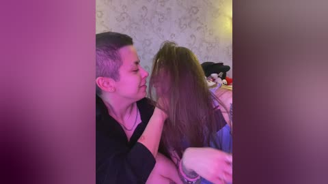 Video of a young man and woman kissing passionately in a dimly lit room with a floral wallpaper background. The woman has long, wavy brown hair, and the man has short, dark hair.