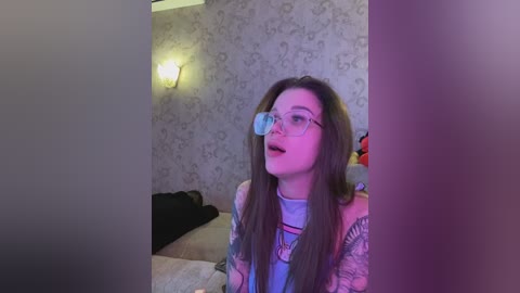 Video of a young woman with long brown hair, wearing glasses, a colorful graphic tee, and tattoos, sitting on a bed with floral wallpaper and a lit lamp in a dimly lit room.