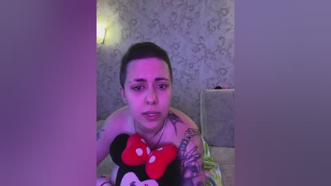 Video of a tattooed woman with short dark hair, holding a Mickey Mouse plush toy, in a dimly lit room with floral wallpaper and a light fixture.