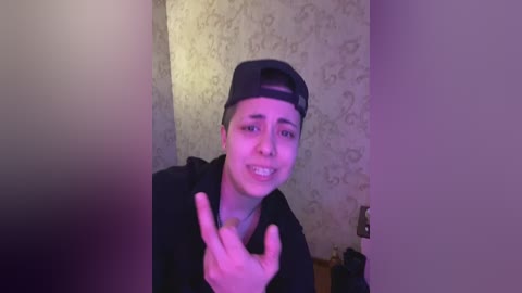 A video of a young man with a light complexion, wearing a black backward baseball cap and a black hoodie, grinning with braces, in a room with textured wallpaper and a purple light.