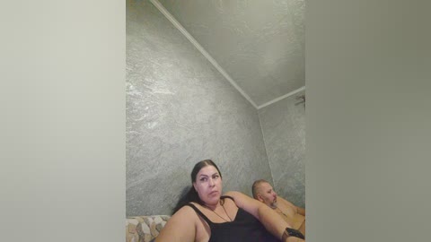 Video of a young, plus-sized woman with dark hair in a black tank top, sitting on a floral-patterned couch. Behind her, a bald man lies on the floor. The room has textured gray walls and a white ceiling.