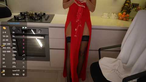 A video of a woman in a red, sheer, high-slit gown with black thigh-high stockings, standing in a modern kitchen.