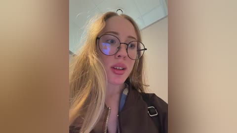 A video of a young, fair-skinned, blonde woman with glasses and a brown jacket, smiling slightly, taken indoors with a ceiling light visible.