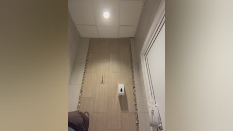 Video of a narrow, beige-tiled hallway with a white ceiling and a round light fixture. A white wall-mounted dispenser is centrally located, with a brown chair and coat rack partially visible in the foreground.