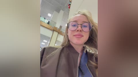 Video of a blonde woman with glasses and a brown jacket, smiling, taken indoors with a blurred background featuring a ceiling and glass partitions.