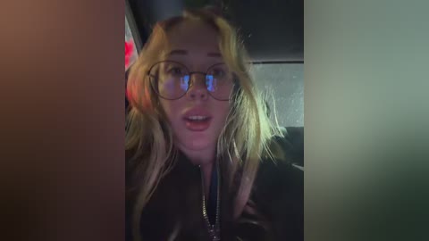 Video of a blonde woman with glasses and a black leather jacket, smiling, with her hand in her hair. Background shows a dimly lit interior.