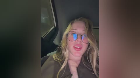 A video of a blonde woman with glasses, smiling, wearing a black coat, sitting in a car at night.