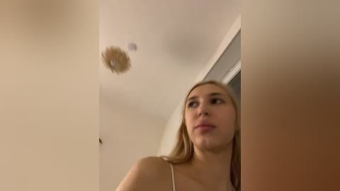A video of a young woman with light skin and straight blonde hair, wearing a white spaghetti strap top, looking upward at a ceiling light fixture. The background is a plain, beige wall with a doorframe.