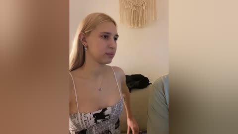 A video of a young woman with fair skin and straight, blonde hair, wearing a black and white patterned spaghetti-strap top, sitting on a beige couch, next to a black bag and a macram\u00e9 wall hanging.