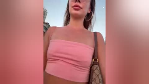 Video of a young woman with fair skin, wearing a pink bandeau top, hoop earrings, and a beige handbag. She has long brown hair and glasses. Background shows an urban skyline.